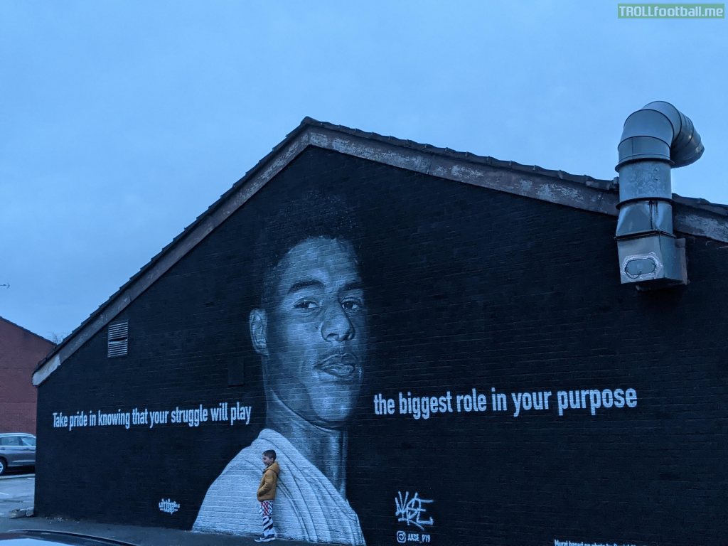 A young boy standing in the shadow of a mural to his hero. Withington, Manchester, UK.